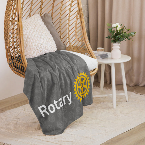 Grey Rotary sherpa blanket draped over a chair in a cozy room, displaying the large Rotary logo and Mark of Excellence motif.