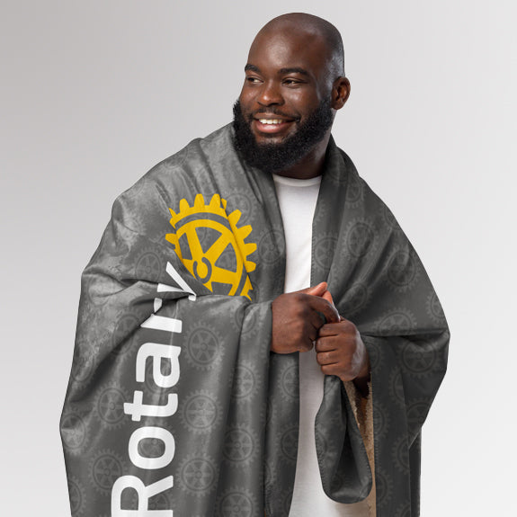 Smiling man wrapped in the grey Rotary sherpa blanket featuring the simplified Masterbrand logo and all-over Mark of Excellence pattern.