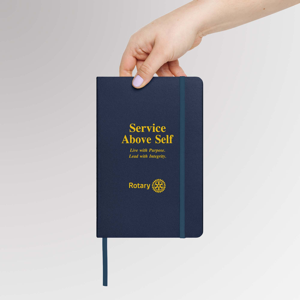 Hand holding a navy blue Rotary notebook featuring gold foil text “Service Above Self” with the Rotary International Masterbrand logo on the front cover, elastic band, and ribbon marker visible.