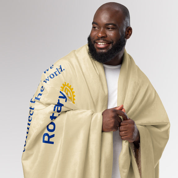 Man smiling while wrapped in the cream Rotary sherpa blanket, partially showing the world map and “Together We Connect the World” text.