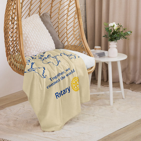 Cream Rotary sherpa blanket casually draped over a chair, showing partial world map design and the “Together We Connect the World” quote.