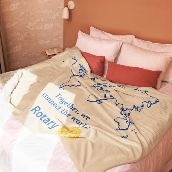 Rotary sherpa blanket fully spread on a bed, displaying the blue world map, simplified Masterbrand logo, and “Together We Connect the World” phrase.