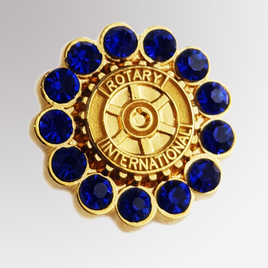 A polished metal pin featuring blue stones and Rotary International's Mark of Excellence logo.